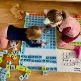 Dwie dziewczynki uczą się czytać, układając litery na planszy. Zabawa pomaga im w nauce jak nauczyć dziecko czytać.