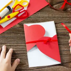 Dziecko tworzy laurkę dla mamy. Na drewnianym stole leżą nożyczki, klej, ołówek, czerwony papier i wstążka.