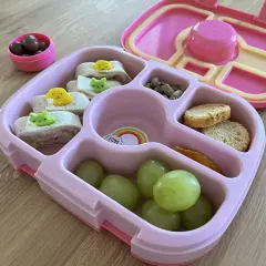 Różowy lunchbox z przekąskami: zawijane tortille z dekoracjami, winogrona, orzeszki pistacjowe, sucharki i czekoladowe drażetki. Idealny pomysł, co do lunchboxa!