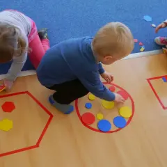 Dzieci układają kolorowe figury geometryczne, tworząc wzory w wyznaczonych obszarach.