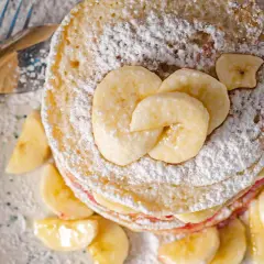 Stos placuszków bananowych dla dzieci, posypany cukrem pudrem i udekorowany plasterkami banana. Obok leżą sztućce.