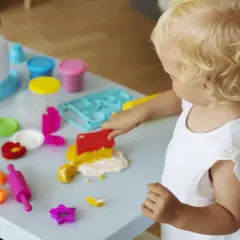 Małe dziecko z blond lokami bawi się masą plastyczną. Wesołe zabawy z 6 miesięcznym dzieckiem przy stoliku pełnym kolorowych foremek i wałków.