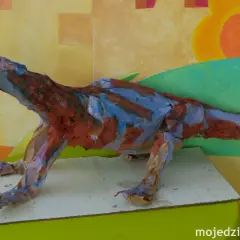 Kolorowy dinozaur praca plastyczna z papieru, z otwartą paszczą i ostrymi zębami, na tle malowanej ściany.