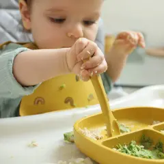 Maluch próbuje samodzielnie jeść, od kiedy masło dla niemowlaka jest na talerzyku. Rączki ubrudzone jedzeniem, ale radość z odkrywania smaków bezcenna.