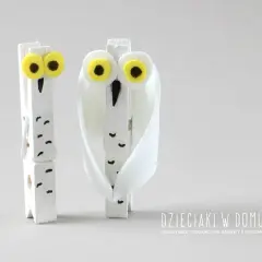 Dwie urocze sowy z papieru, wykonane z drewnianych klamerek, z wielkimi żółtymi oczami i białymi piórami.