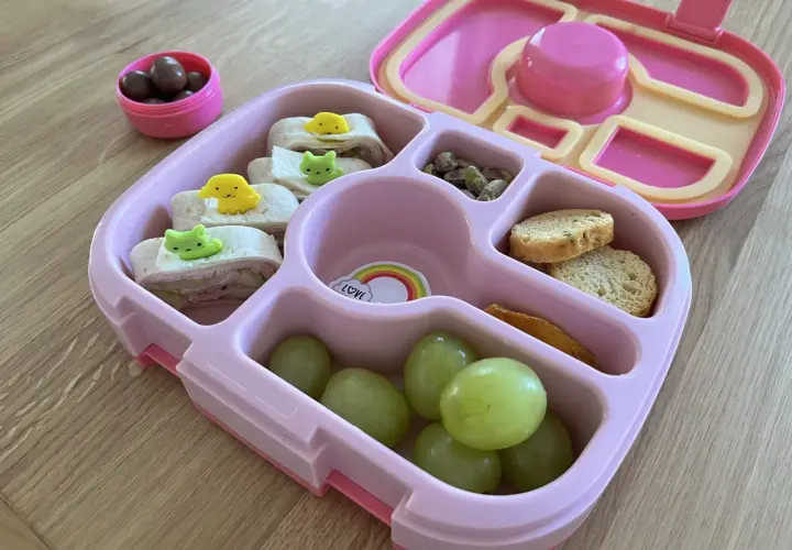 Różowy lunchbox z przekąskami: zawijane tortille z dekoracjami, winogrona, orzeszki pistacjowe, sucharki i czekoladowe drażetki. Idealny pomysł, co do lunchboxa!