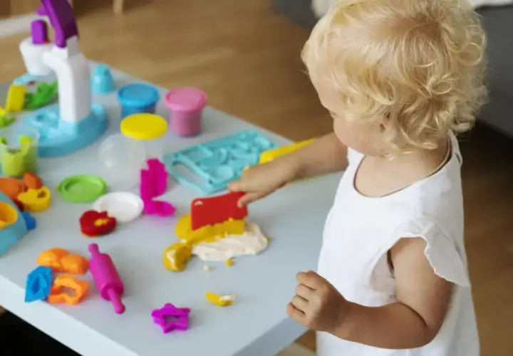 Małe dziecko z blond lokami bawi się masą plastyczną. Wesołe zabawy z 6 miesięcznym dzieckiem przy stoliku pełnym kolorowych foremek i wałków.