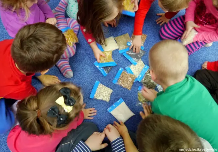 Dzieci siedzą w kółku na niebieskim dywanie, badając zawartość woreczków z różnymi produktami. To wspaniałe zabawy sensoryczne!