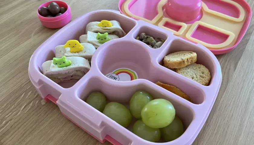 Różowy lunchbox z przekąskami: zawijane tortille z dekoracjami, winogrona, orzeszki pistacjowe, sucharki i czekoladowe drażetki. Idealny pomysł, co do lunchboxa!