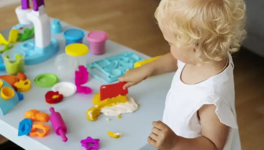 Małe dziecko z blond lokami bawi się masą plastyczną. Wesołe zabawy z 6 miesięcznym dzieckiem przy stoliku pełnym kolorowych foremek i wałków.