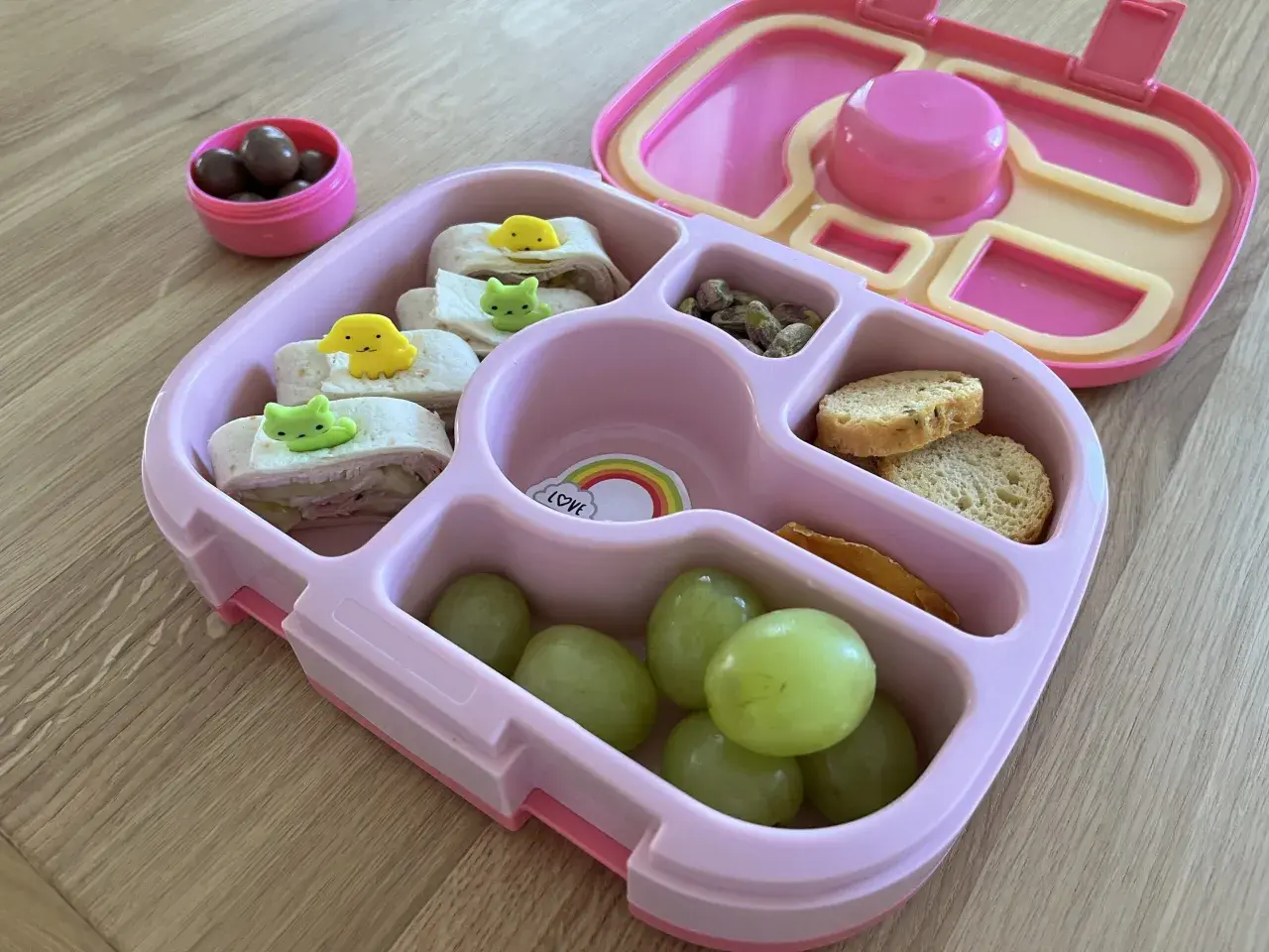 Różowy lunchbox z przekąskami: zawijane tortille z dekoracjami, winogrona, orzeszki pistacjowe, sucharki i czekoladowe drażetki. Idealny pomysł, co do lunchboxa!
