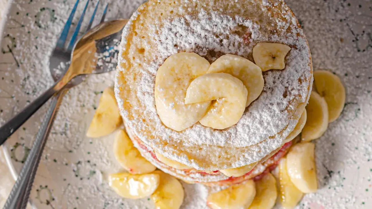 Stos placuszków bananowych dla dzieci, posypany cukrem pudrem i udekorowany plasterkami banana. Obok leżą sztućce.