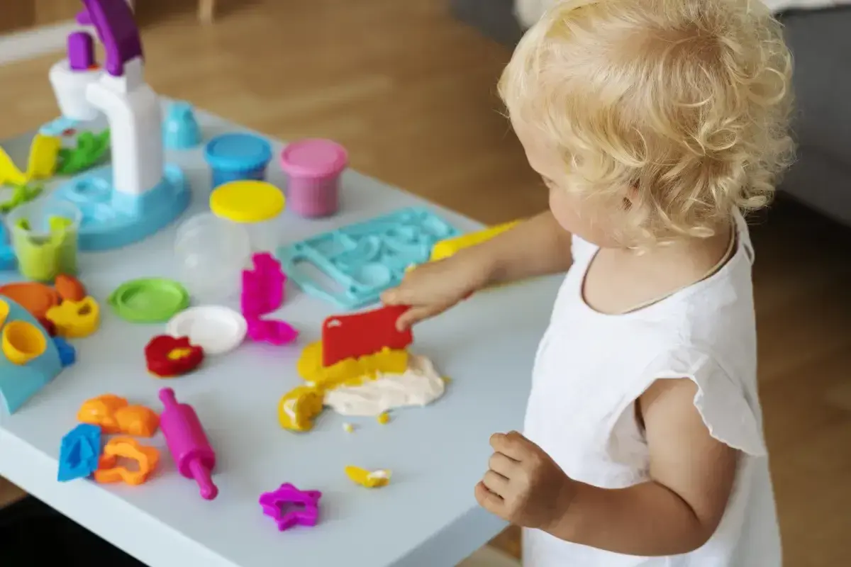 Małe dziecko z blond lokami bawi się masą plastyczną. Wesołe zabawy z 6 miesięcznym dzieckiem przy stoliku pełnym kolorowych foremek i wałków.