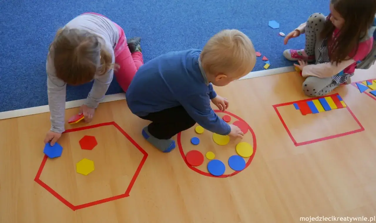 Dzieci w przedszkolu bawią się kolorowymi figurami geometrycznymi, tworząc wzory na podłodze.