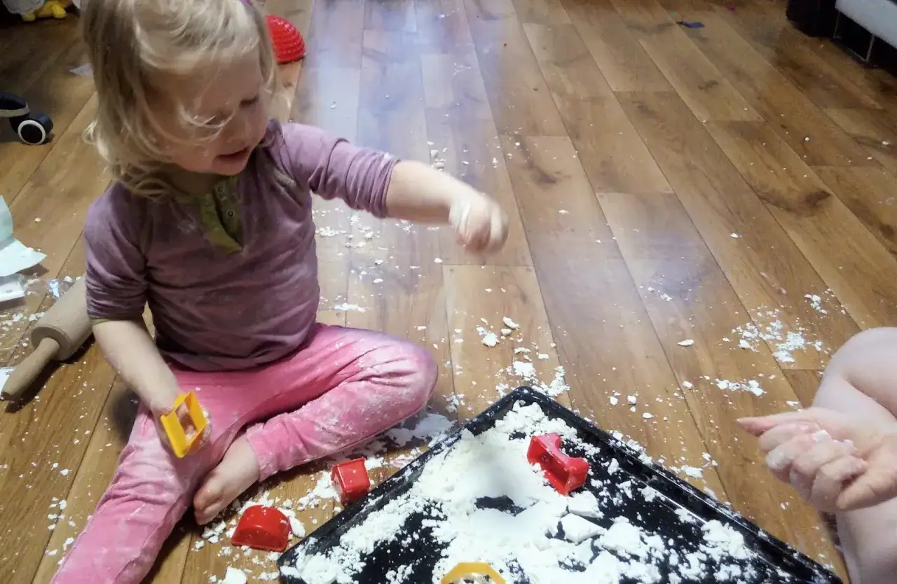 Dziecko bawi się w tworzenie sztucznego śniegu z mąki i wody, rozsypując go na drewnianej podłodze.